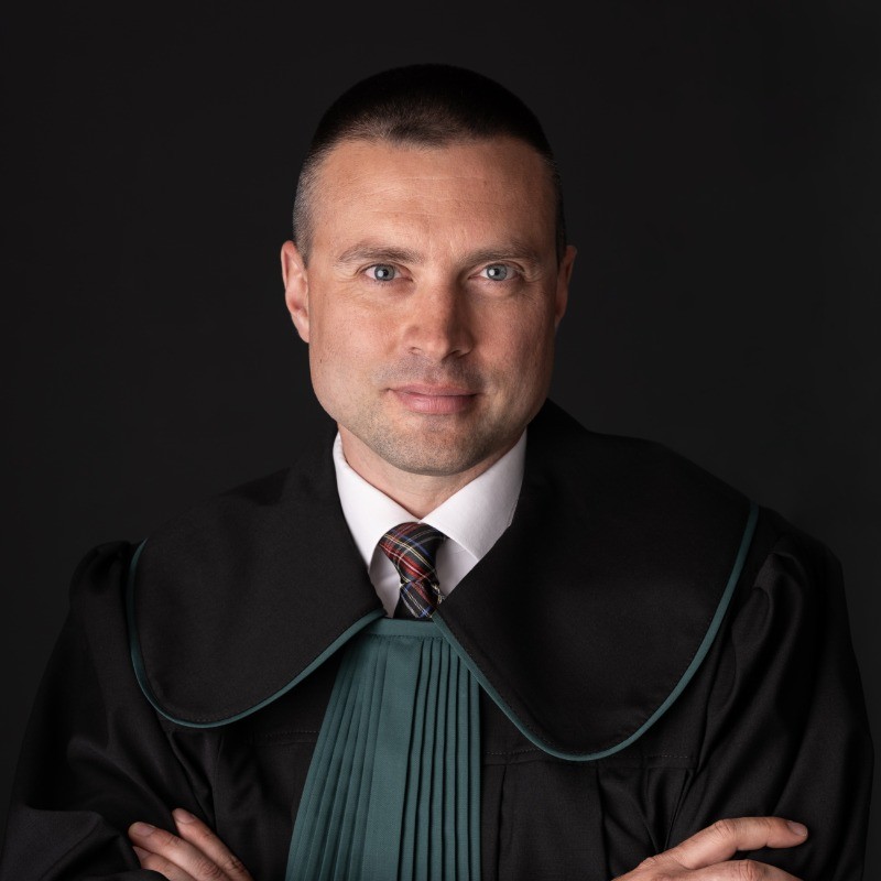 Professional headshot of Oskar Luty in formal legal attire with black court robes and green collar trim